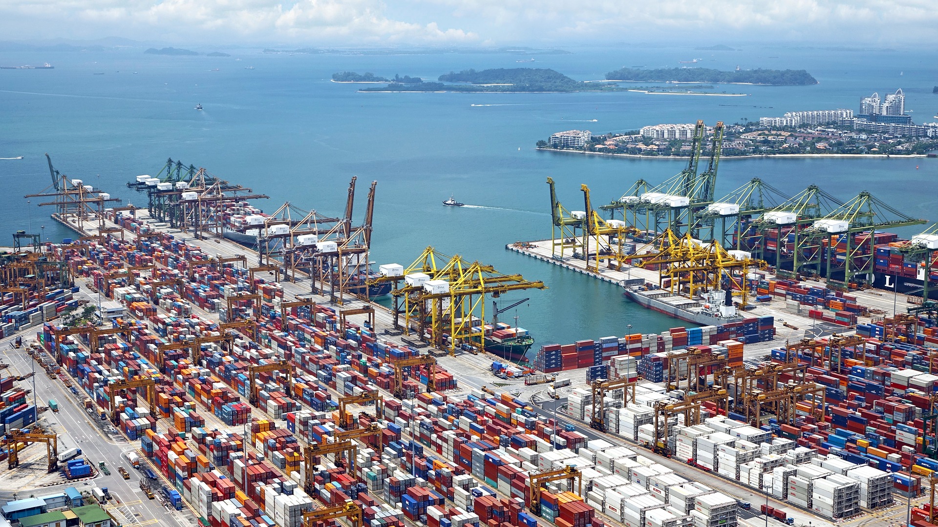 A high-angle aerial view of a sprawling and busy commercial seaport. Thousands of brightly colored shipping containers are densely stacked across the docks, while massive yellow and green gantry cranes load cargo onto large ships moored in the bright blue harbor.