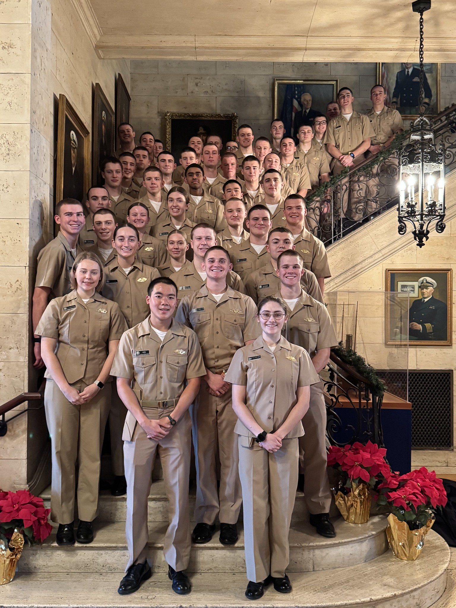 A large group of smiling midshipmen in short-sleeved khaki uniforms posing together for a photo on a grand indoor marble staircase. The elegant setting features an ornate wrought-iron railing, framed portraits hanging on the stone walls, and festive red poinsettias at the base of the steps.