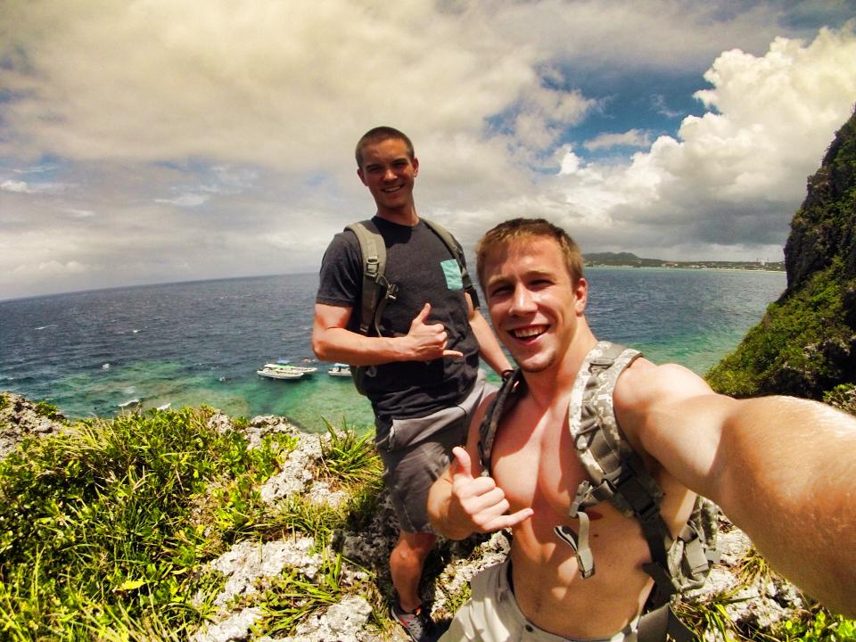 Midshipmen Yeik and Lindgren take a smiling selfie while hiking on a lush, rocky cliffside during liberty in Okinawa, Japan. Both are making the 'shaka' hand sign, with a beautiful view of the clear turquoise ocean and small boats down below.