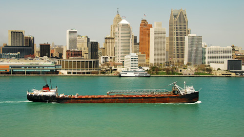 A long, traditional Great Lakes bulk carrier ship sailing down a vibrant green waterway, with the dense Detroit city skyline towering in the background.