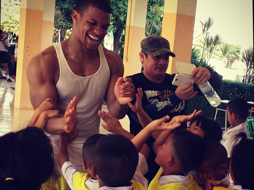 Midshipman Mickel McGann smiles broadly while giving high-fives to a group of young school children wearing yellow vests during a volunteer visit to the Pena School in Thailand. Another man stands next to him, taking a picture of the joyful moment with a smartphone.