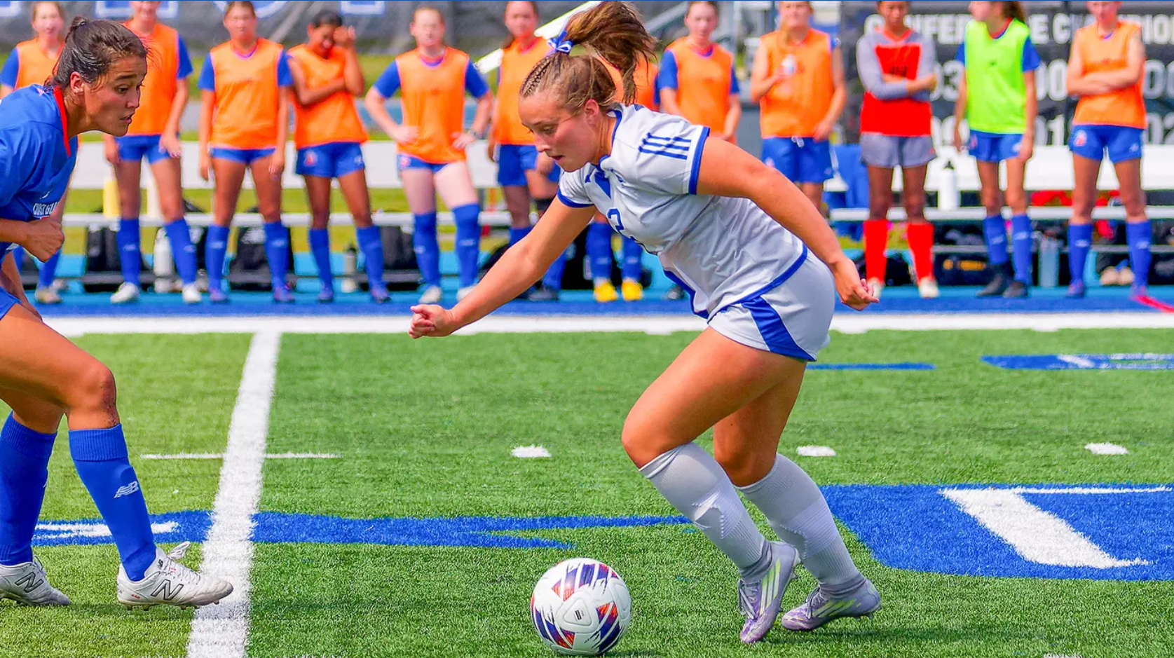 A female soccer player in a white and blue uniform dribbles a soccer ball down a green turf field during a match. She is being guarded closely by an opposing player in blue on the left, while teammates in colorful practice pinnies watch from the sidelines in the blurred background.