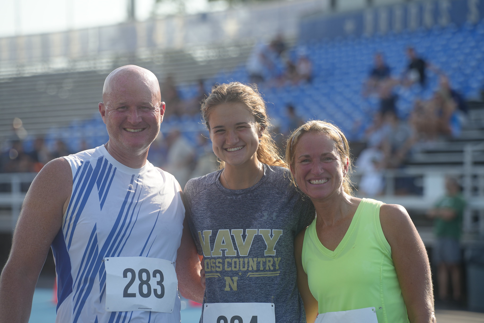 Three smiling participants pose together for a close-up photo on a blue track at the Peter J. Rackett ’61 Memorial Run. The man and two women are wearing athletic clothing and race bibs, sharing a cheerful moment with stadium bleachers visible in the background.