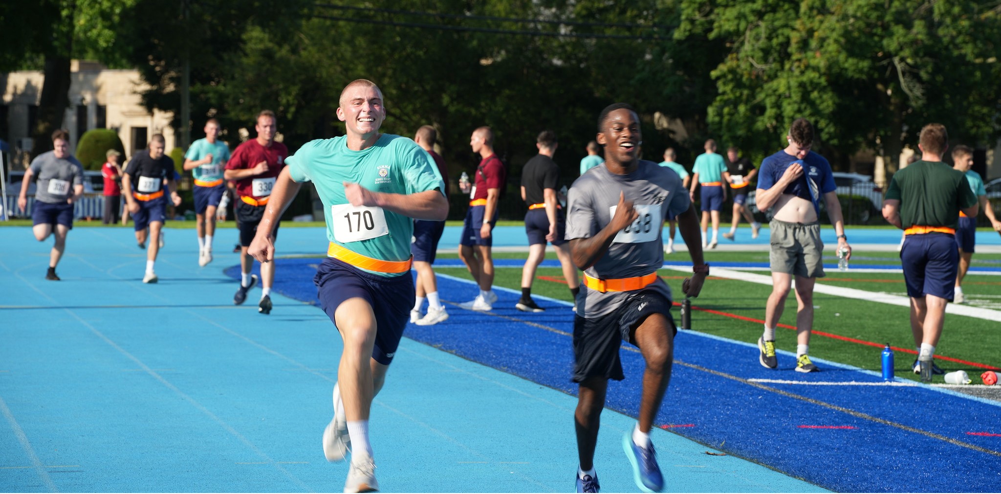 Two smiling midshipmen sprint side-by-side down a bright blue track during the Peter J. Rackett ’61 Memorial Run. They are wearing physical training gear, orange reflective belts, and race bibs, with a large group of fellow participants running behind them on a sunny day.