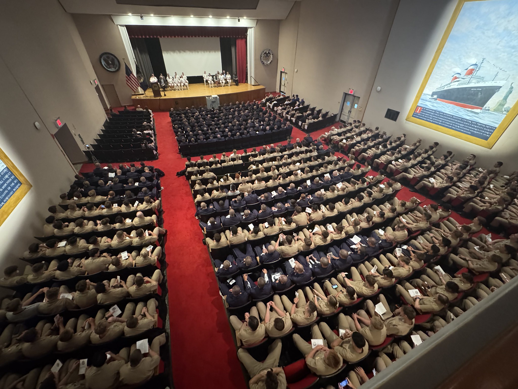 A high-angle, wide shot of a packed auditorium during the USMMA Academic Convocation. Hundreds of midshipmen, mostly in khaki uniforms with a front section in dark blue uniforms, are seated in rows facing a stage. Faculty and leadership are seated on the well-lit stage near a wooden podium and an American flag. Bright red carpet lines the wide aisles, and a massive maritime mural of a ship passing the Statue of Liberty hangs on the right wall.