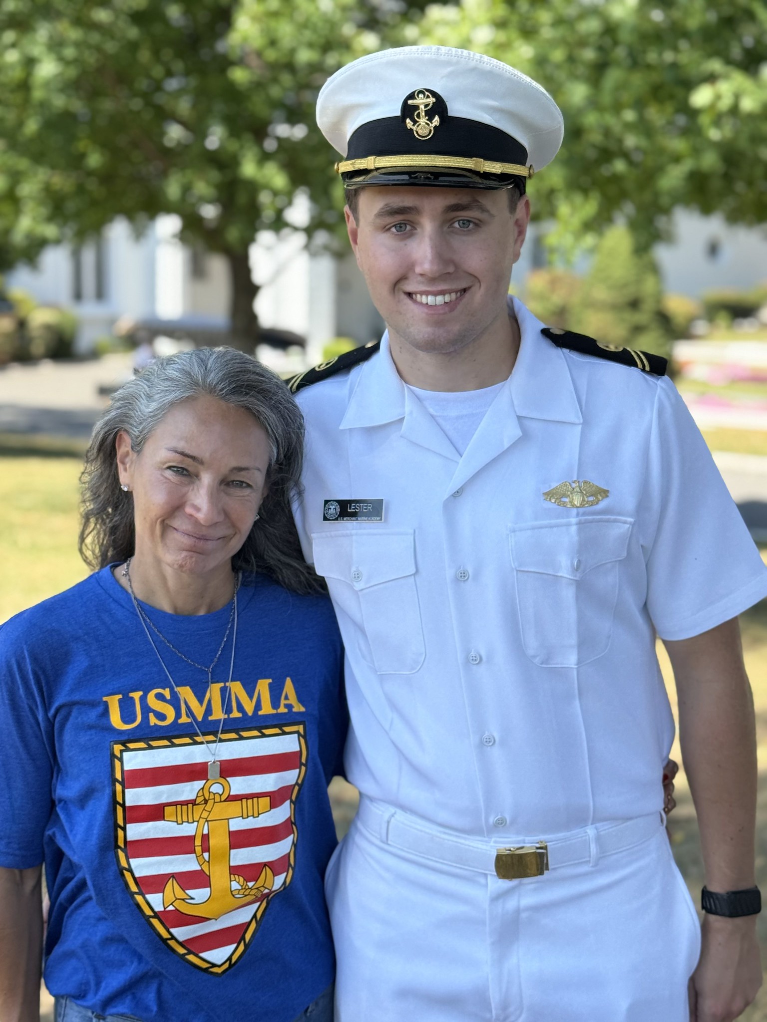 A waist-up portrait of a USMMA midshipman and a woman, likely his parent, standing proudly together outdoors on a sunny day during Parents' Weekend. The smiling midshipman is wearing his crisp Summer White uniform, complete with a combination cover, shoulder boards, and a nametag reading "Lester." Beside him, the woman smiles warmly, wearing a bright blue t-shirt featuring the USMMA shield and anchor logo. The background is softly blurred, displaying green trees and a glimpse of a white campus building.