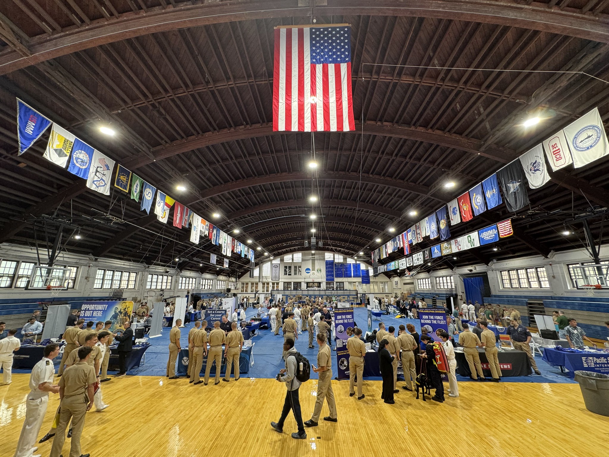 A wide, elevated view of the Sea Fair career event inside the wood-ceilinged O’Hara Hall. A large American flag and colorful pennants hang above. Below, dozens of USMMA midshipmen in Service Khaki uniforms network at vendor booths and promotional tables set up across the gym floor.