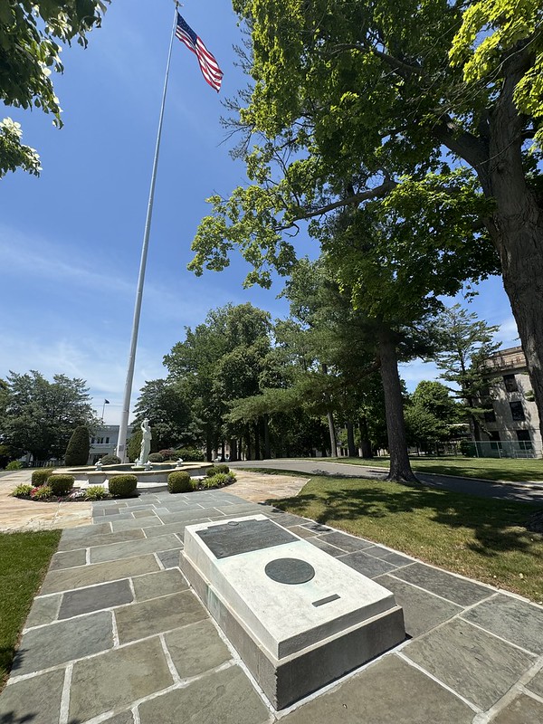 A scenic, sunny view of a memorial area on the USMMA campus. In the foreground, a low stone monument with dark plaques rests on a paved slate walkway. The path leads toward a circular stone fountain featuring a central statue, surrounded by manicured hedges. Beside the fountain, a tall white flagpole proudly flies the American flag against a clear blue sky. Lush, mature trees frame the right side of the scene, casting shade over the grass, with glimpses of campus buildings visible in the background.