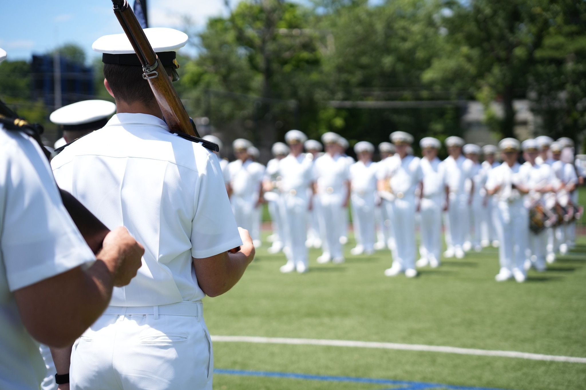 An over-the-shoulder perspective of a midshipman in a Summer White uniform standing with a ceremonial rifle resting on his shoulder. In the background across the green turf field, a large formation of midshipmen, including members of the Regimental Band holding brass instruments, stand uniformly at attention under a bright, sunny sky.