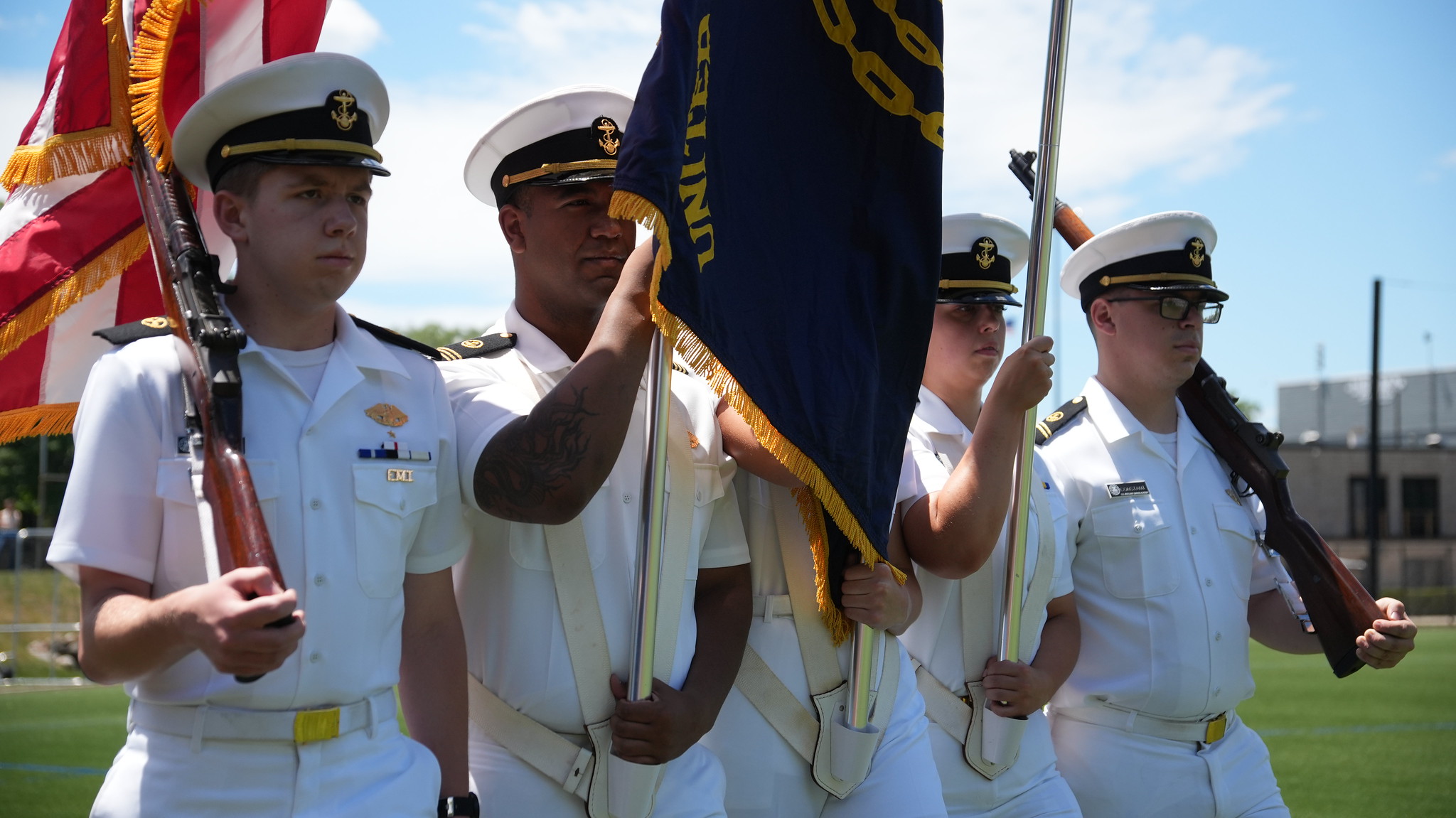 A close-up shot of the USMMA color guard marching in formation during a Regimental Parade. Four midshipmen wearing crisp Summer White uniforms are marching across a green turf field. The two outer midshipmen are carrying ceremonial rifles resting on their shoulders, while the two midshipmen in the center are carrying large, gold-fringed flags supported by white harnesses.