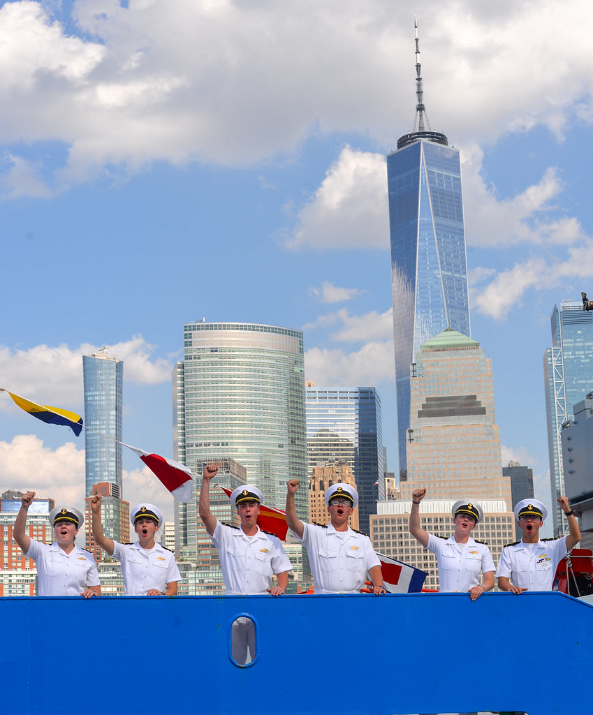 Six USMMA midshipmen in crisp Summer White uniforms stand behind a bright blue ship's railing, cheering enthusiastically with their right fists raised in the air. Colorful nautical signal flags are strung in the background. Behind the midshipmen, the Lower Manhattan skyline rises majestically, prominently featuring One World Trade Center towering under a bright blue sky with scattered clouds.