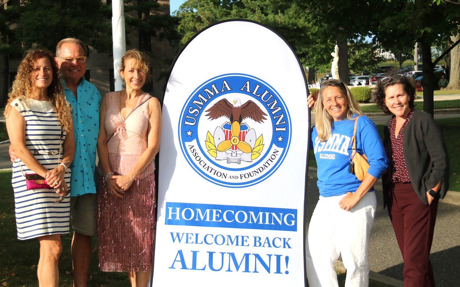 Five people—four women and one man—smile as they pose outdoors next to a large white pop-up banner on a sunny campus. The banner features the USMMA Alumni Association and Foundation logo and reads, "HOMECOMING WELCOME BACK ALUMNI!" in bold blue lettering. The group is dressed in casual weekend attire, with one woman proudly wearing a blue "USMMA Alumni" sweatshirt. Trees, a paved walkway, and parked cars are visible in the background.