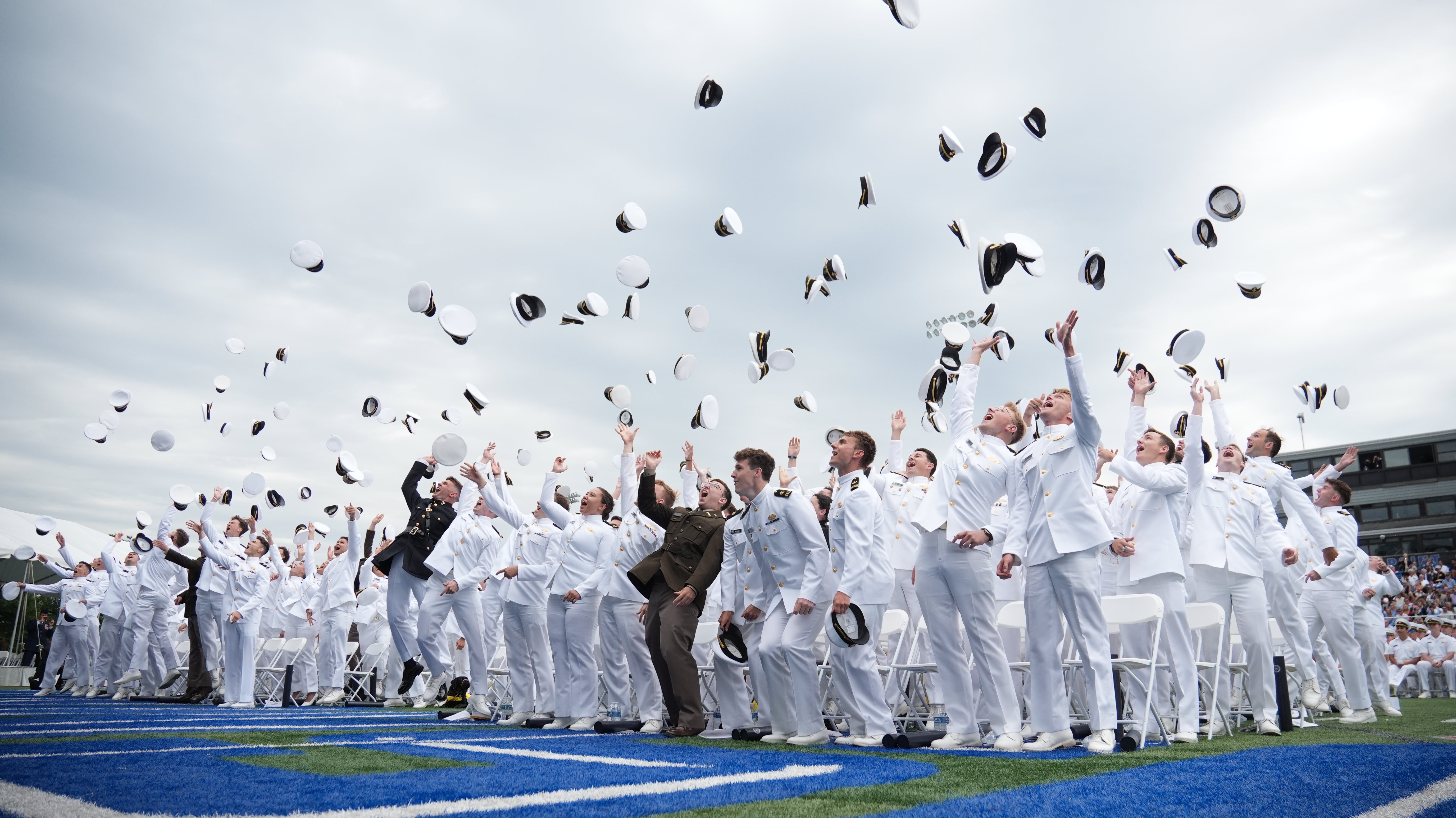 A large group of joyous United States Merchant Marine Academy graduates in dress white and military uniforms celebrating their commencement by tossing their white caps high into the air. The graduates are cheering with their arms raised, standing on a blue turf field with white folding chairs and stadium seating visible in the background under an overcast sky.
