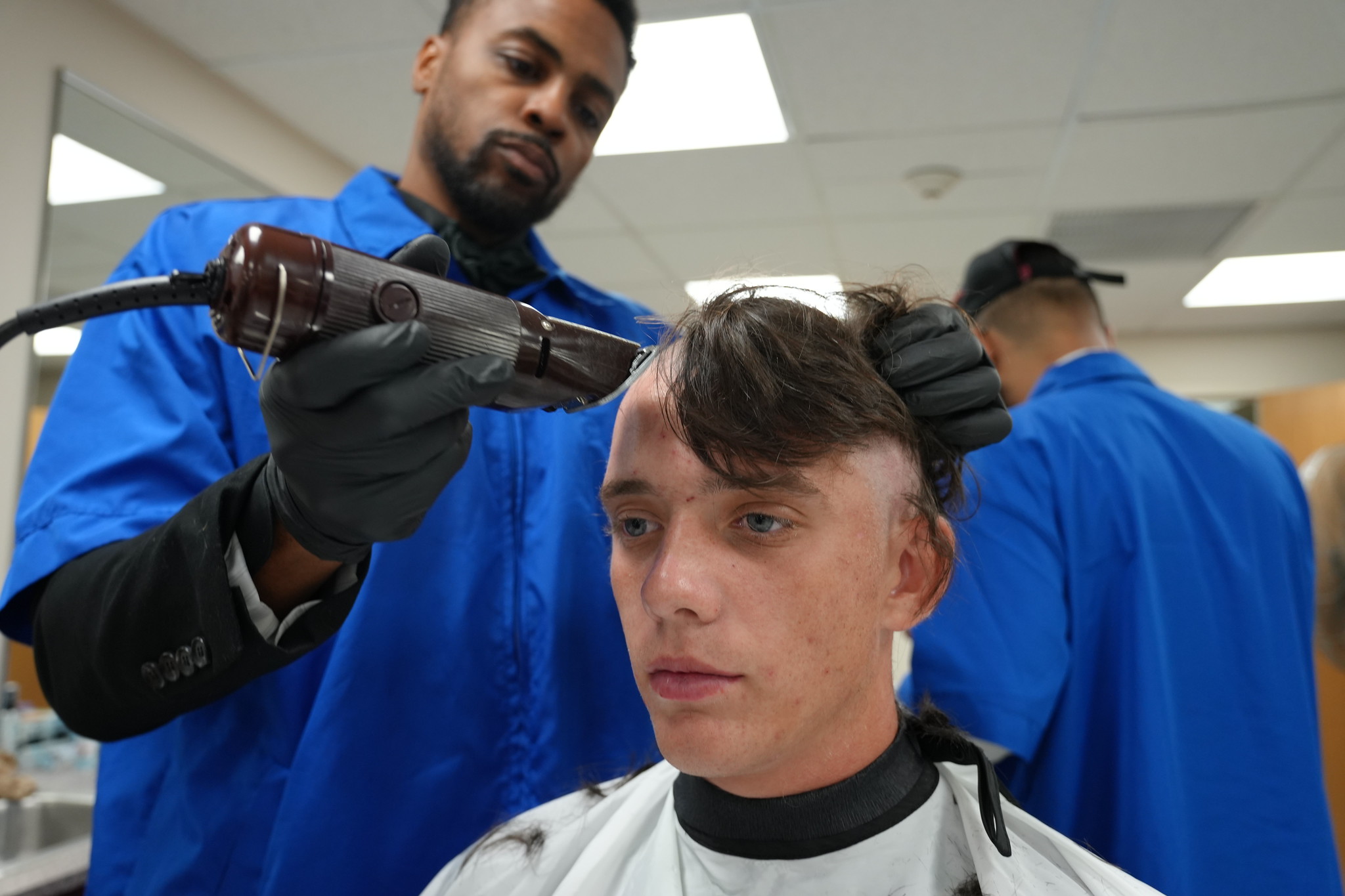 A young midshipman candidate stares straight ahead with a stoic expression while a barber wearing a blue smock and black gloves uses electric clippers to shave his head during Indoctrination. Half of the candidate's hair has already been buzzed to the scalp, and another barber is visible working in the bright background.