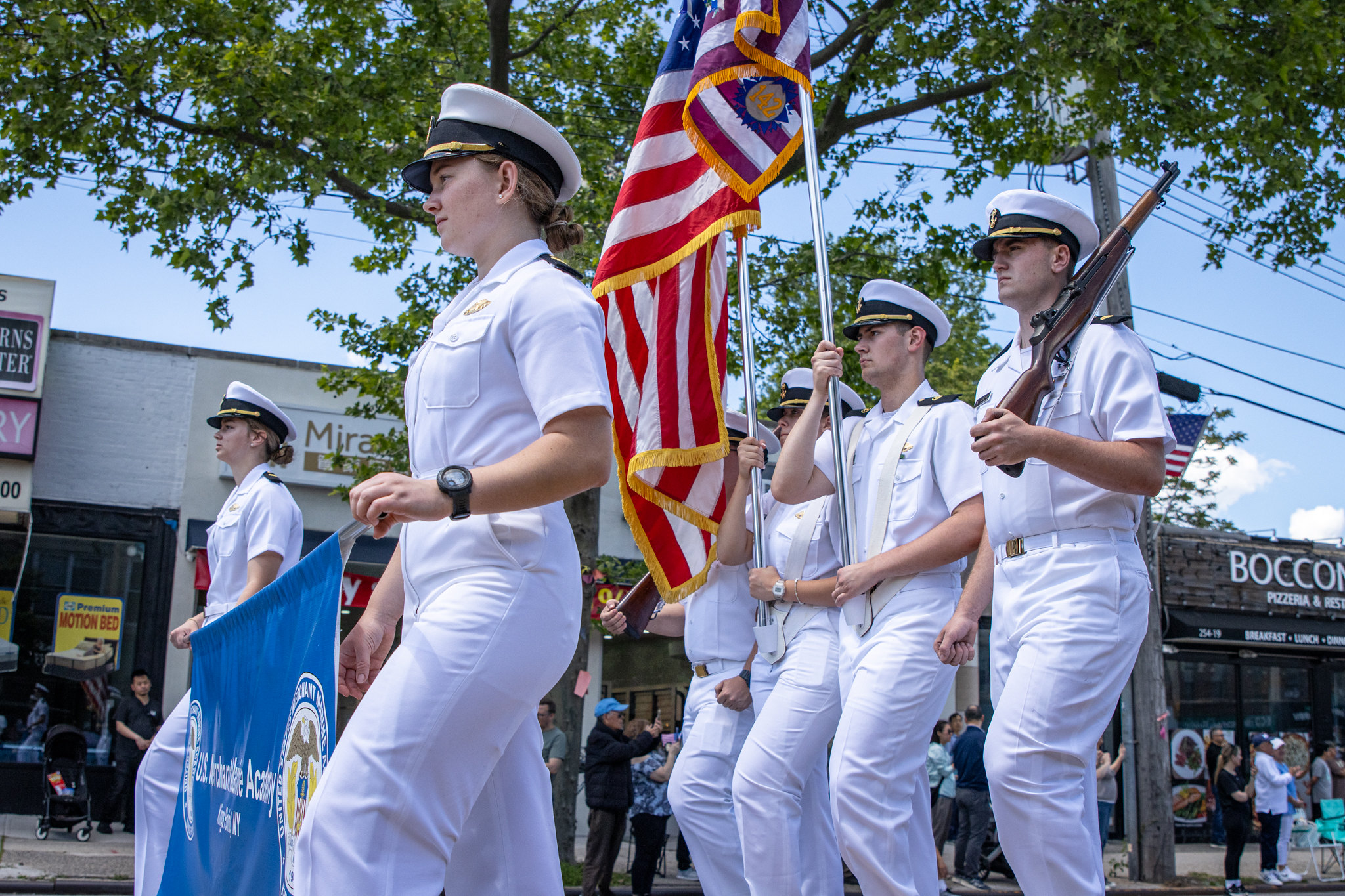 A group of U.S. Merchant Marine Academy cadets in crisp white uniforms march in a parade down a city street. They are carrying a blue academy banner, the American flag, a unit flag, and ceremonial rifles while spectators watch from the sidewalks.