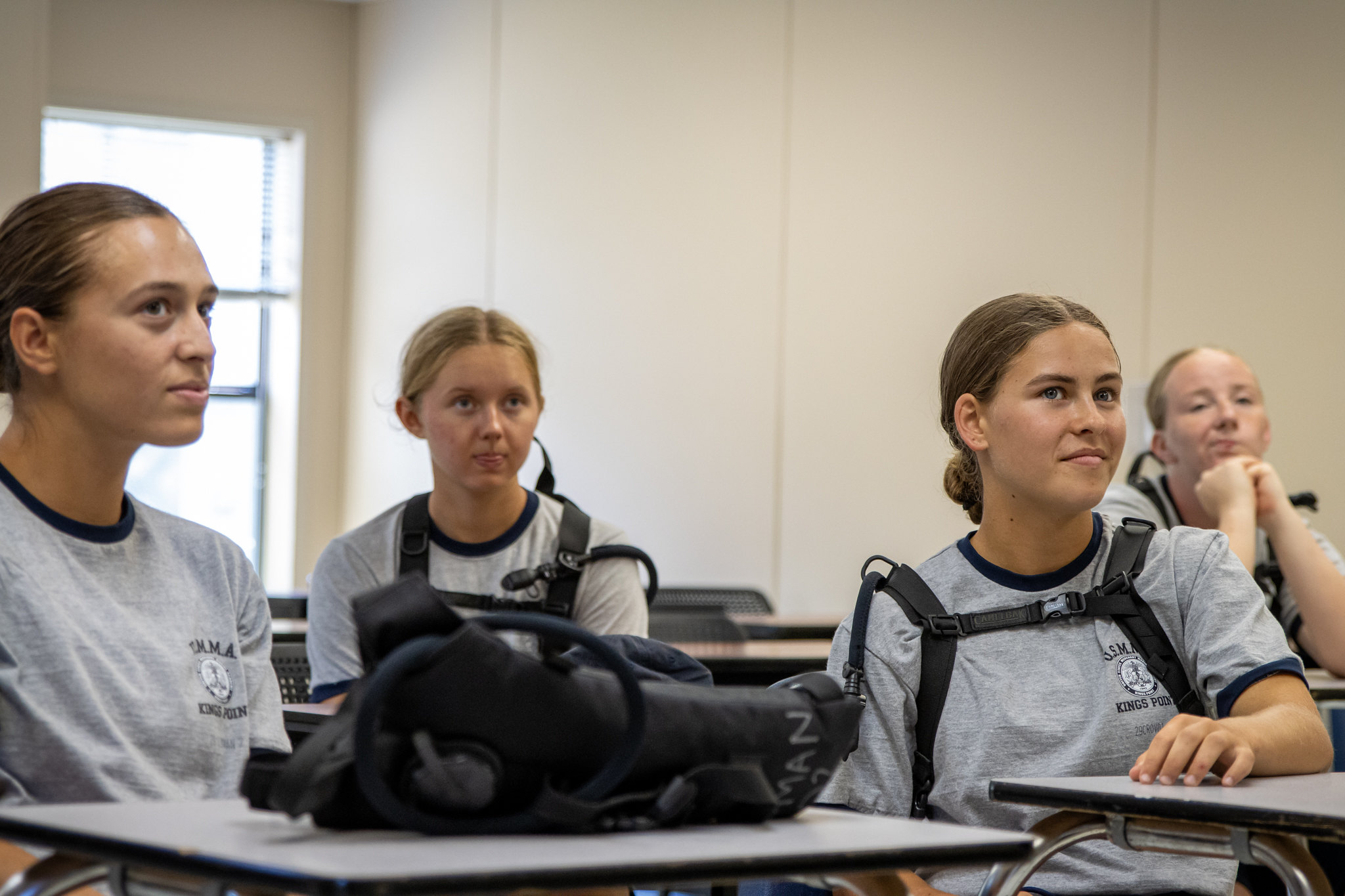 Four young women in matching grey Kings Point t-shirts sit attentively at desks in a classroom, looking forward toward an unseen speaker. Several of the students are wearing black hydration backpacks with drinking tubes over their shoulders.