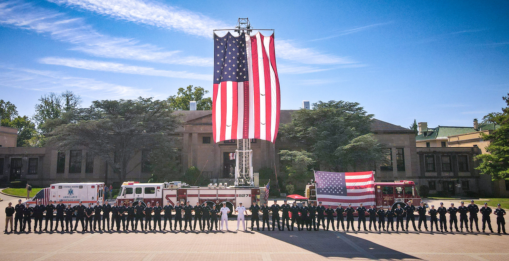 A long line of uniformed personnel stands in front of an ambulance and two fire engines parked outside a large stone building. A massive American flag hangs vertically from the extended ladder of the center fire engine, with a second flag draped over the side of the vehicle on the right.