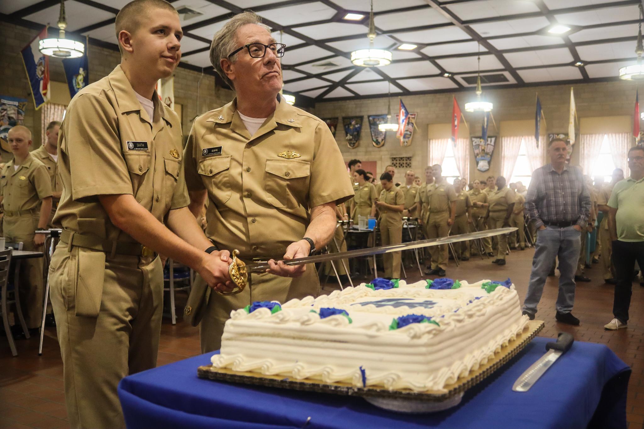 A young man and an older man in matching khaki military uniforms jointly hold a ceremonial sword to cut a large sheet cake. They are standing in a large, flag-decorated dining hall, surrounded by a crowd of onlookers in similar uniforms watching the ceremony.