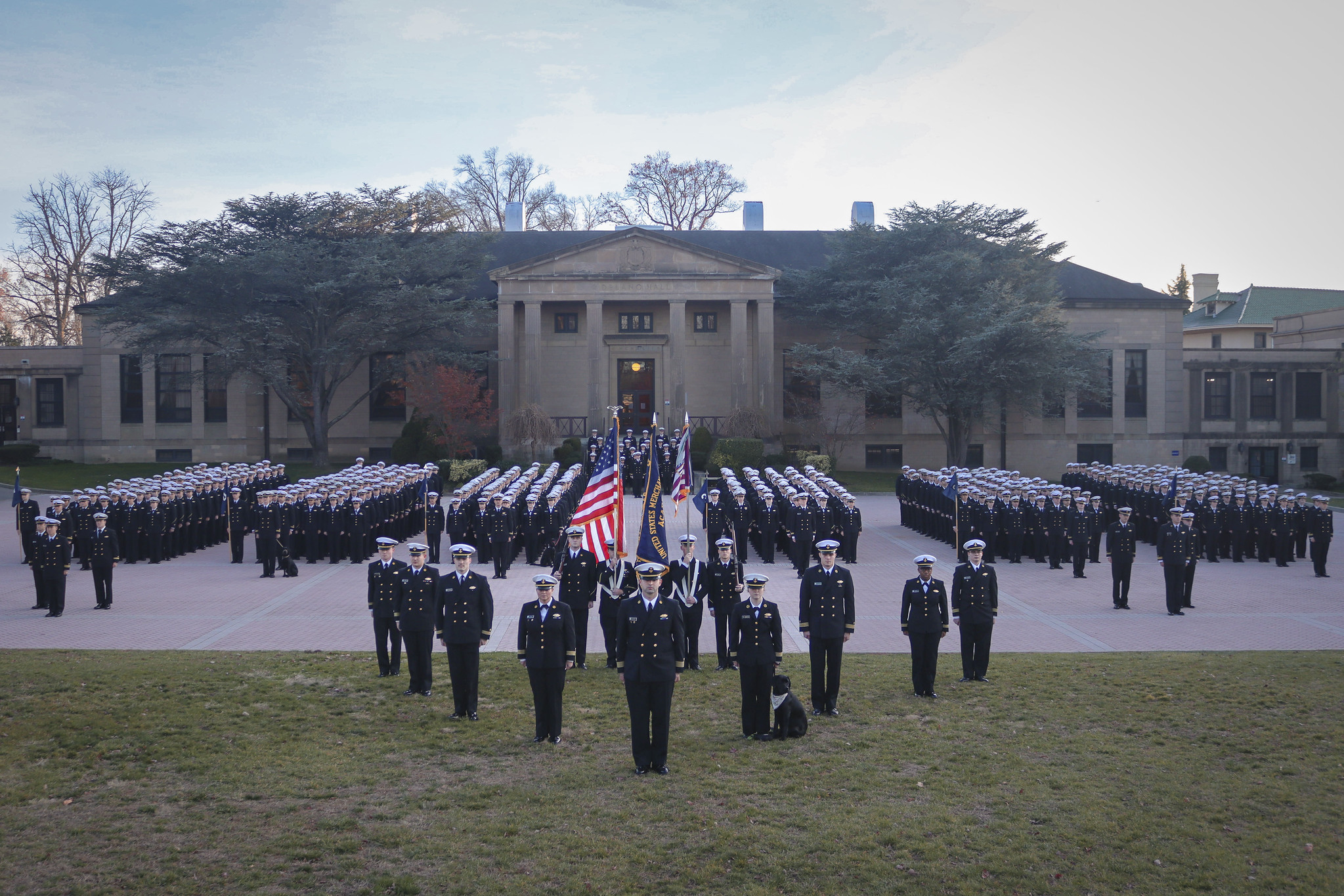 A large formation of uniformed military personnel stands in precise rows on a brick courtyard in front of a grand stone building with columns. In the foreground, a smaller group of leaders and a color guard holding the American flag stand on the grass, accompanied by a black dog sitting beside one of the officers.