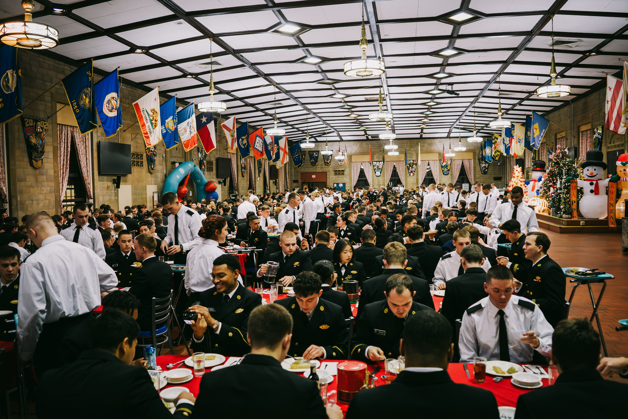 A large, crowded dining hall is filled with people in military-style uniforms eating at tables covered with red tablecloths. State flags hang along the walls beneath a patterned ceiling, and festive holiday decorations, including an inflatable snowman, are visible in the background.
