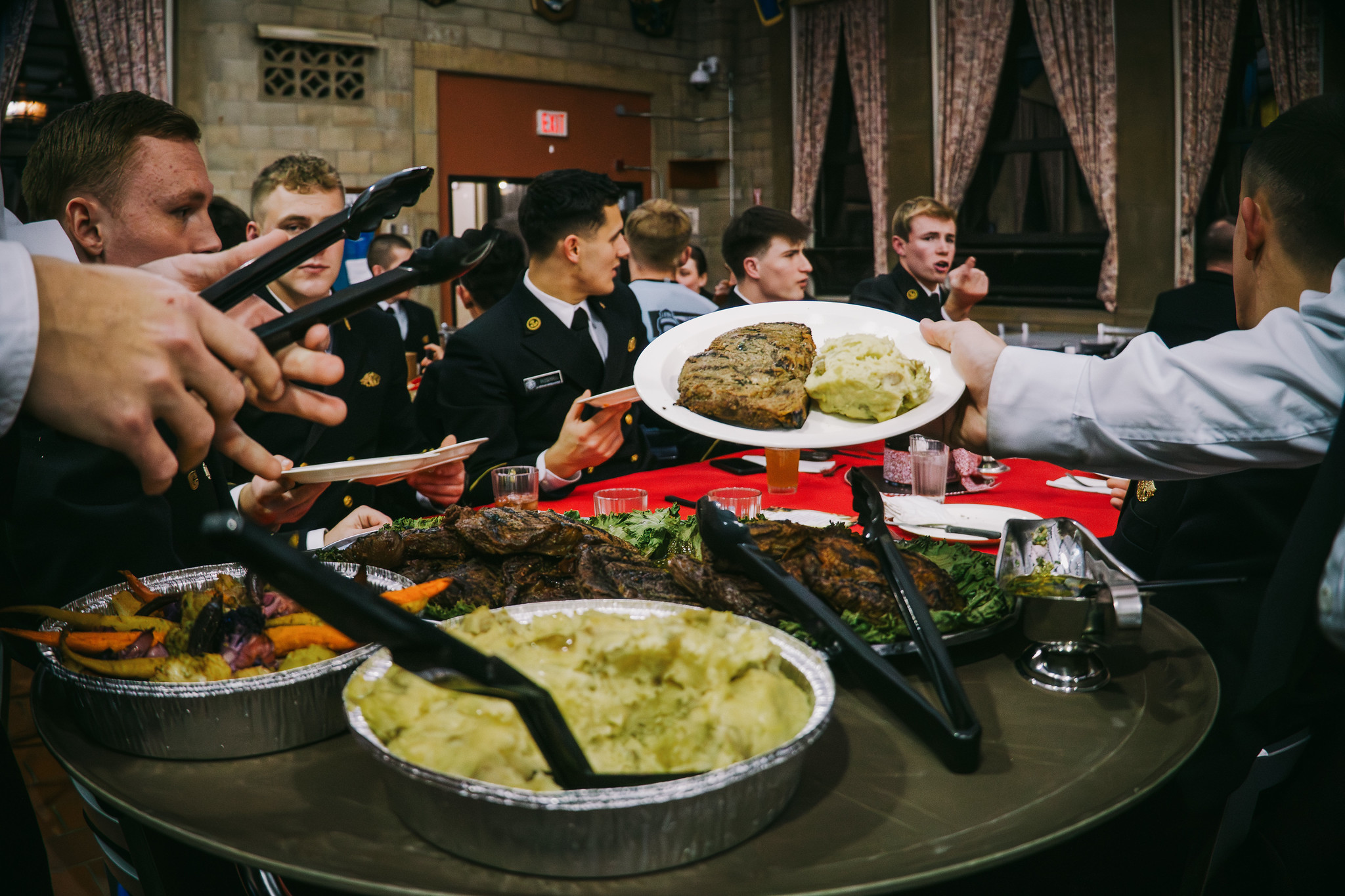 Young men in dark military-style uniforms are seated at a banquet table with a red tablecloth in a formal dining hall. In the foreground, a person hands out a freshly plated meal of steak and mashed potatoes over a serving table filled with large foil trays of catered food.