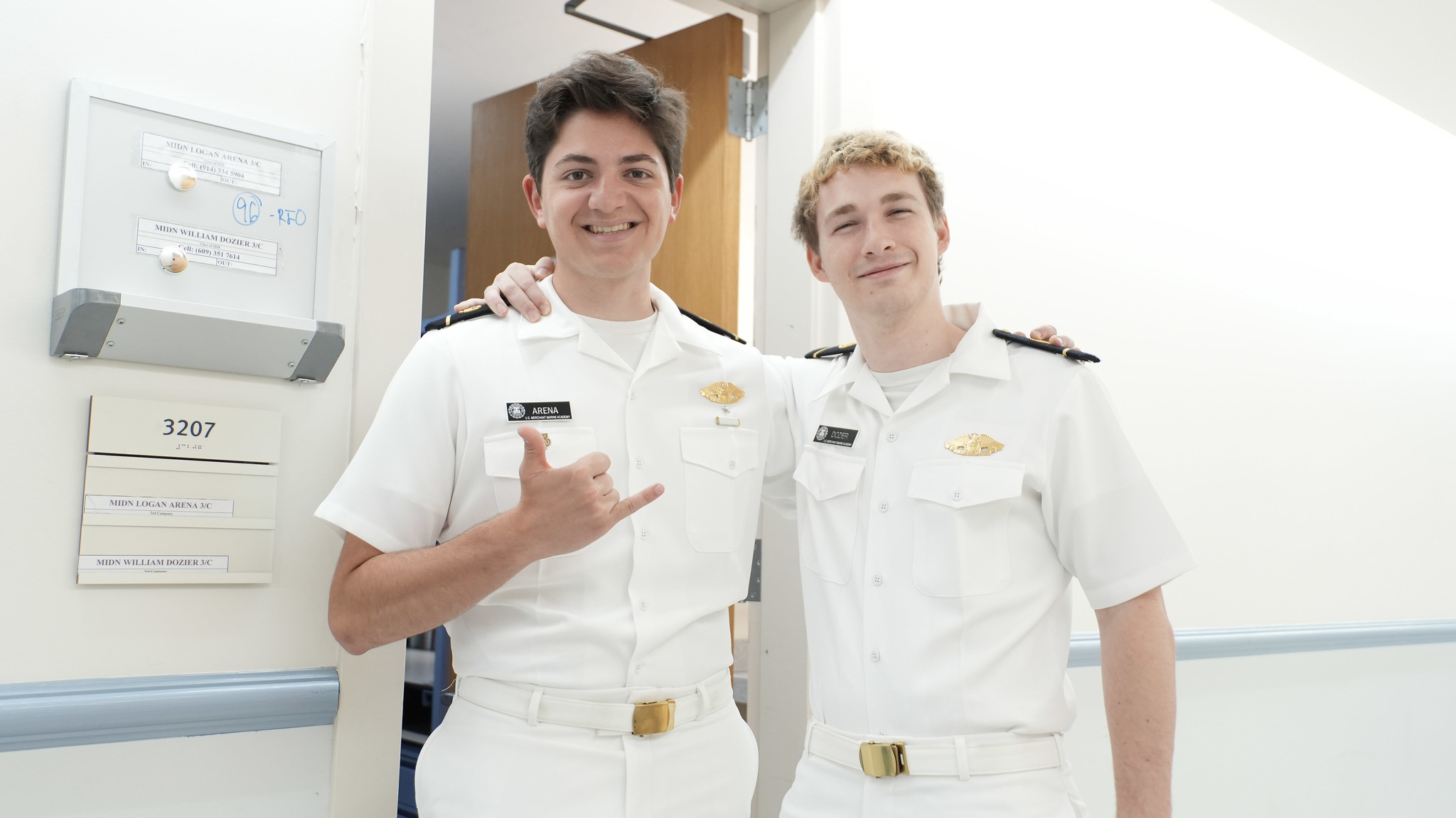 Two male midshipmen in Summer White uniforms pose together, smiling in a bright barracks hallway outside room 3207. Standing with arms around each other's shoulders, the midshipman on the left proudly makes a "shaka" hand sign. A placard on the wall next to the open door identifies them as Third Class Midshipmen Logan Arena and William Dozier.