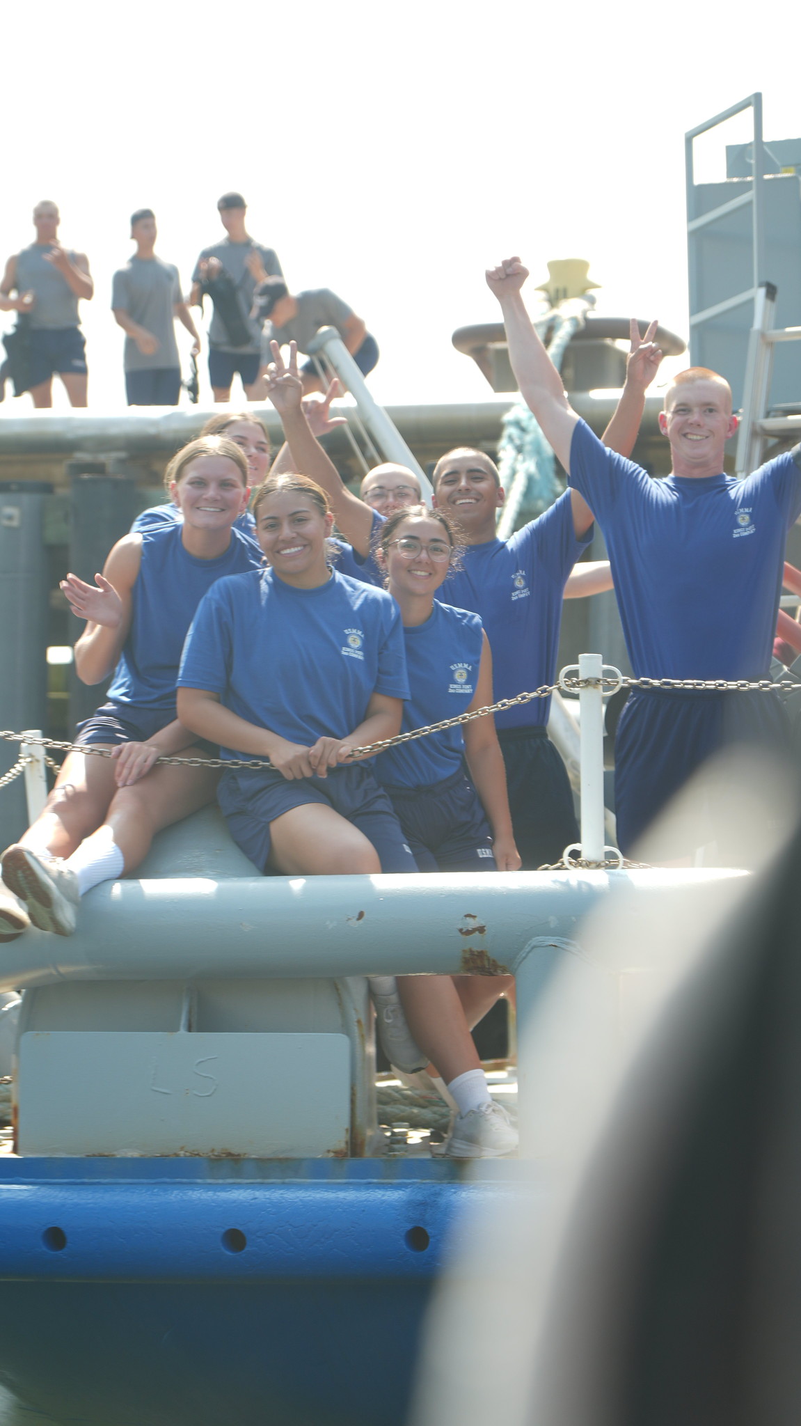 A group of smiling young adults in matching blue uniforms pose excitedly on a large mooring bollard, waving and making peace signs. In the background, other individuals in similar athletic attire stand on a raised ship deck under a bright sky.