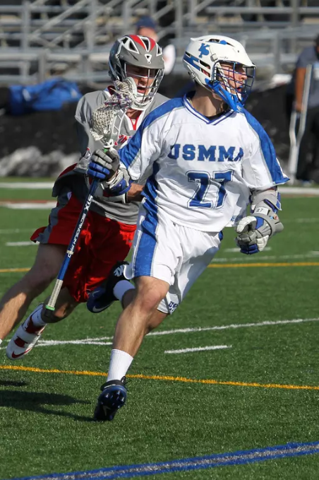 A lacrosse player wearing a white and blue USMMA uniform runs down a turf field during a game. An opposing player in a grey and red uniform chases closely behind him, holding a lacrosse stick.