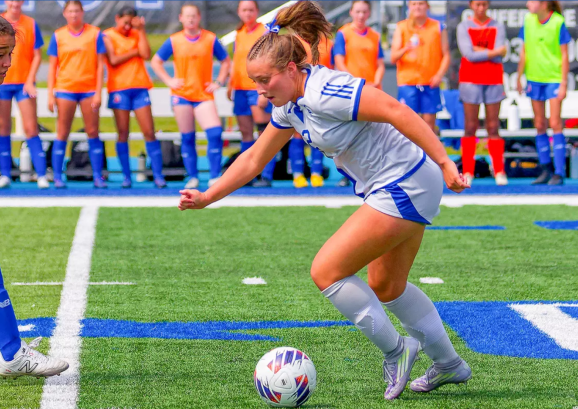 A female soccer player in a white and blue uniform dribbles a soccer ball down a green turf field during a match. She is being guarded closely by an opposing player in blue on the left, while teammates in colorful practice pinnies watch from the sidelines in the blurred background.