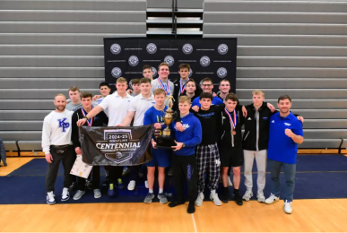 A large group photograph of the men's wrestling team and coaches celebrating a victory in a gymnasium. The team is gathered in front of a branded backdrop and bleachers, proudly displaying a large gold trophy and holding a black banner that reads '2024-25 Wrestling Centennial Conference Champions'.